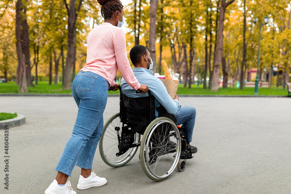 Young black disabled guy in wheelchair holding paper bag with food, returning home from shopping with wife during covid Young black disabled guy in wheelchair holding paper bag with food, returning home from shopping with wife during covid