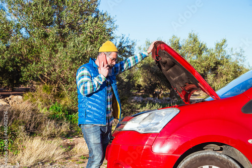 Bearded man talking on the phone next to his broken down car on the road