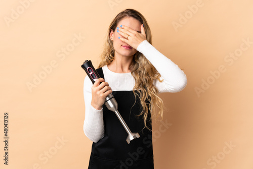 Young brazilian woman using hand blender isolated on beige background covering eyes by hands. Do not want to see something