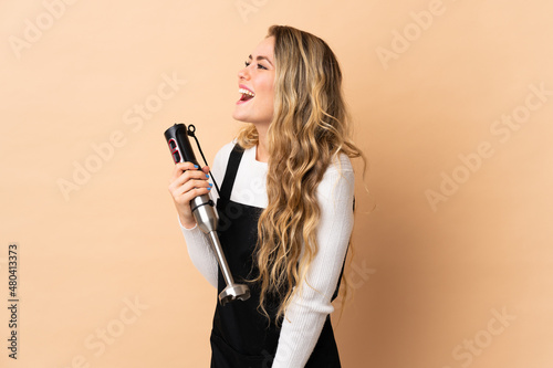 Young brazilian woman using hand blender isolated on beige background laughing in lateral position