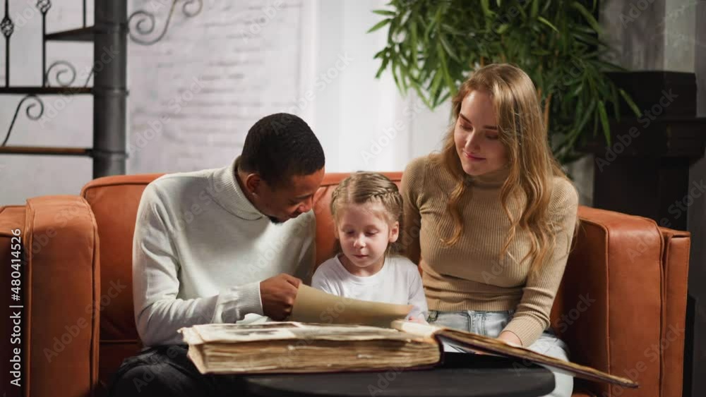African-American man shows photo of grandparents to little girl in album sitting with blonde wife on sofa in cozy living room slow motion African-American man shows photo of grandparents to little girl in album sitting with blonde wife on sofa in cozy living room slow motion