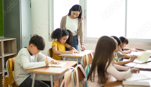 Asian Female teacher helping school girl while her writing in note book in elementary school lessons. Pupils during exam in classrrom. Education knowledge concept