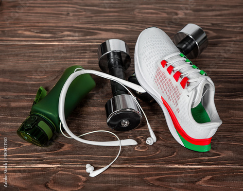 Sports equipment white sneakers  metal dumbbells  a bottle of water and headphones on a wooden background.
