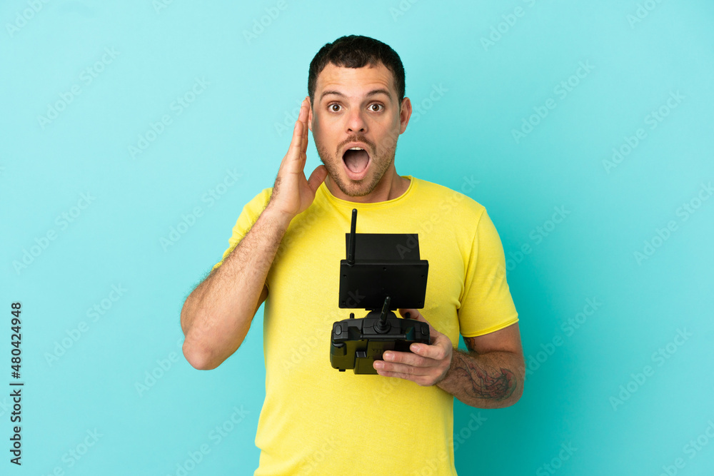 Brazilian man holding a drone remote control over isolated blue background with surprise and shocked facial expression Brazilian man holding a drone remote control over isolated blue background with surprise and shocked facial expression