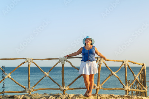 A young brunette woman in a white skirt and a sun hat on the seashore stands against the background of the sea and sky. © finist_4
