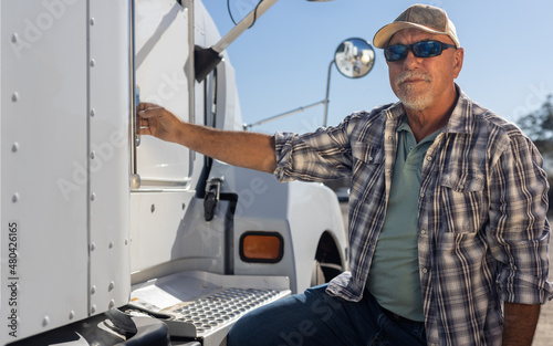 Hispanic middle aged semi truck driver sits next to truck. Experienced senior trucker next to big rig at the job. Portrait of transportation and logistics worker next to equipment on a sunny day.