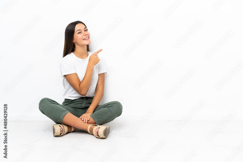 Teenager girl sitting on the floor pointing finger to the side