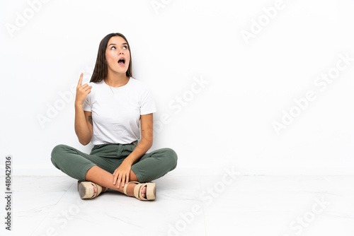Teenager girl sitting on the floor pointing up and surprised