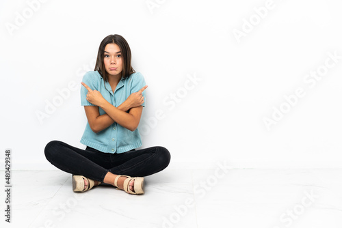 Teenager girl sitting on the floor pointing to the laterals having doubts