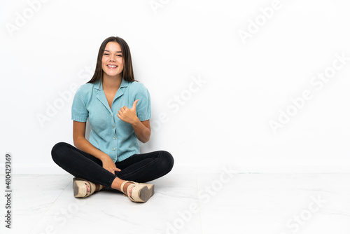 Teenager girl sitting on the floor pointing to the side to present a product