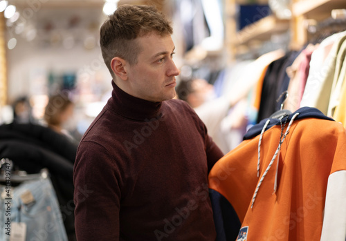 Photo of a handsome brunet young man in a burgundy sweater who chooses clothes in a store in a mall. Shopping concept