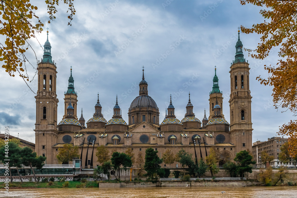 Cathedral-Basilica of Our Lady of the Pillar on the embankment of Ebro river with yellow water in Zaragoza on an autumn day, Spain. Panoramic view of the Spanish Roman Catholic Church