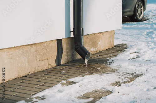 Photo of a downpipe in winter with frozen water and a car in the background.