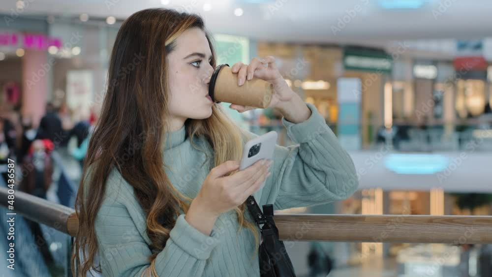 Young caucasian woman millennial girl standing indoors in mall business company looking at mobile phone reading message typing texting on smartphone using wifi app drinking coffee tea from paper cup