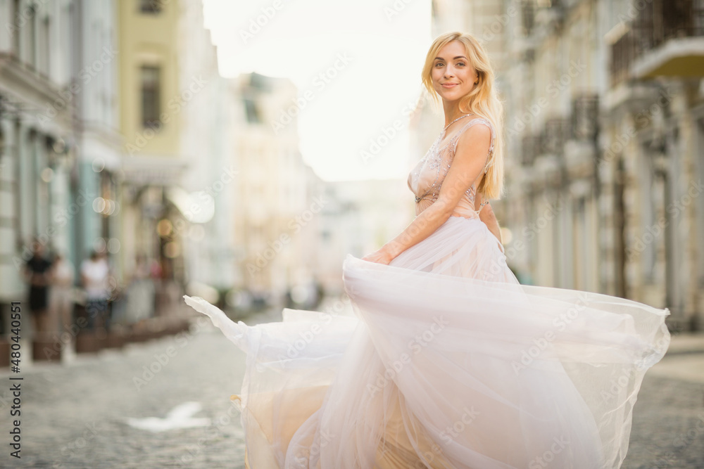 Obraz premium Pretty smiling young woman with long blond hair in elegant flying light dress running along the street