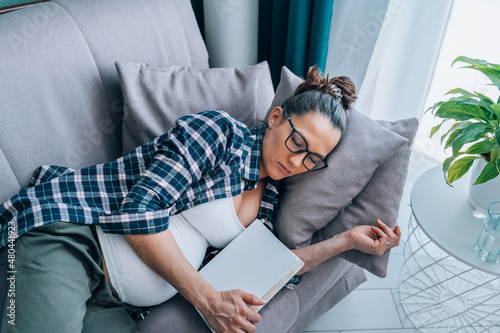 Middle age pregnant woman in eyeglasses wearing checkered shirt sleep lying on grey couch in living room hugging massive book. Daydream, nap, lack of sleeping