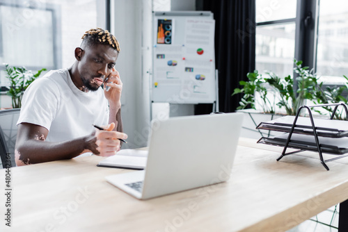 african american man with vitiligo holding pen near notebook and laptop while talking on smartphone in office.