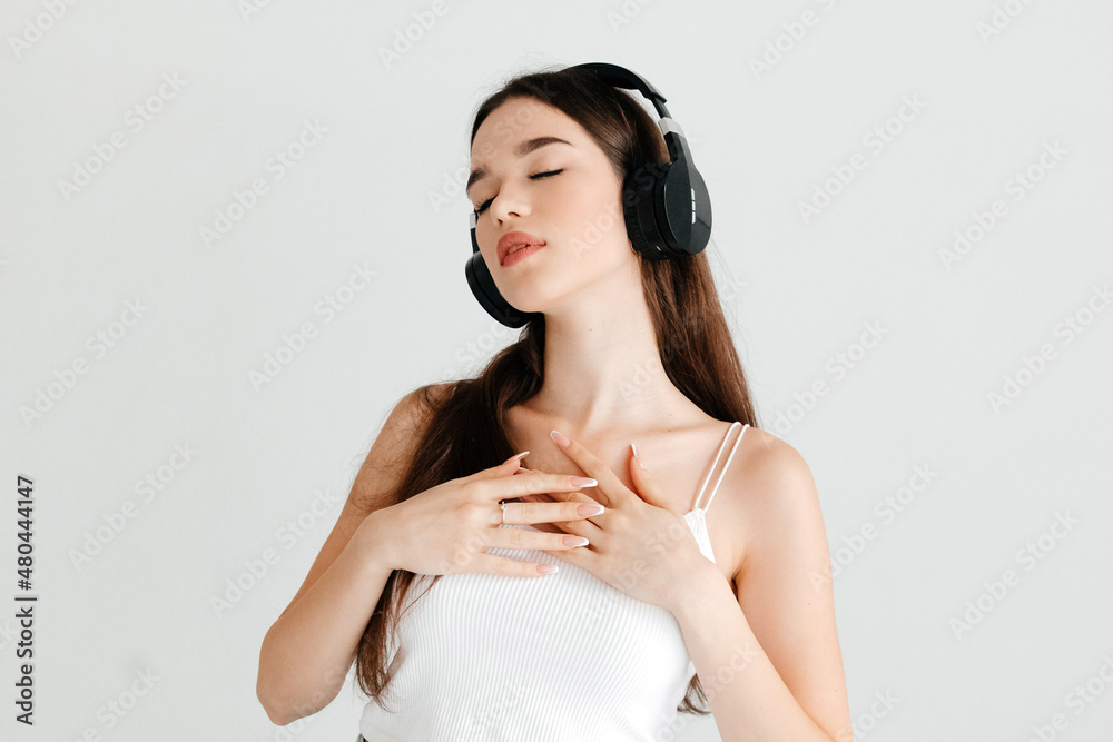 Beautiful young woman with closed eyes, dark hair listening to her favorite song in black wireless headphone, holding her hands on her chest, on white background Beautiful young woman with closed eyes, dark hair listening to her favorite song in black wireless headphone, holding her hands on her chest, on white background
