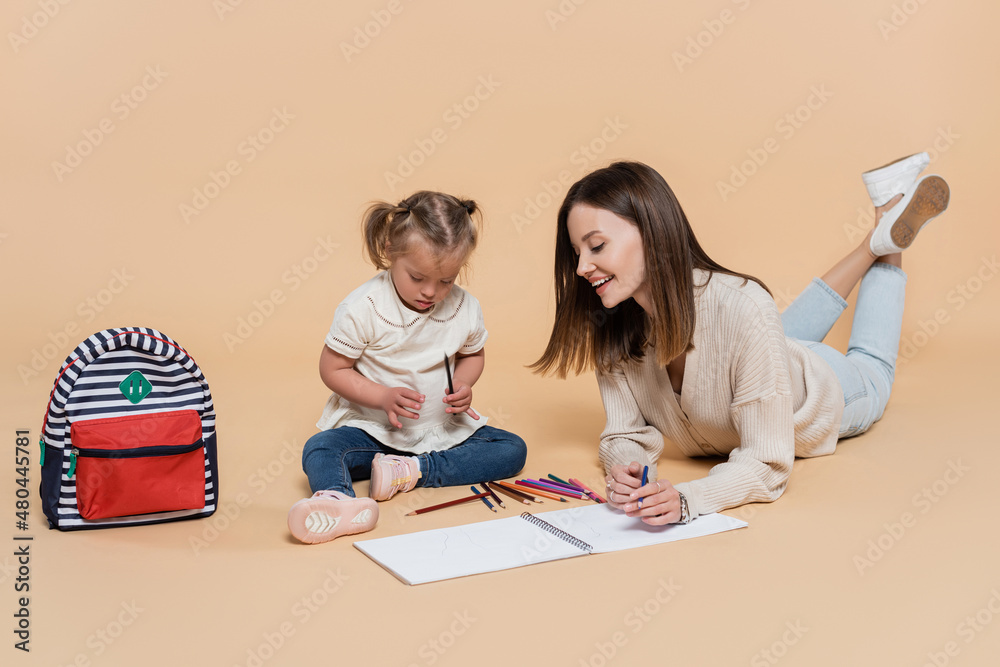 joyful mother lying near kid with down syndrome drawing near colorful pencils and backpack on beige. joyful mother lying near kid with down syndrome drawing near colorful pencils and backpack on beige.