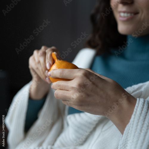 Smiling woman peels juicy ripe sicilian tangerine at home, selective focus on hands. Happy female wrapped in white knitted plaid eating seasonal winter fruits, defocused. High vitamin C food concept