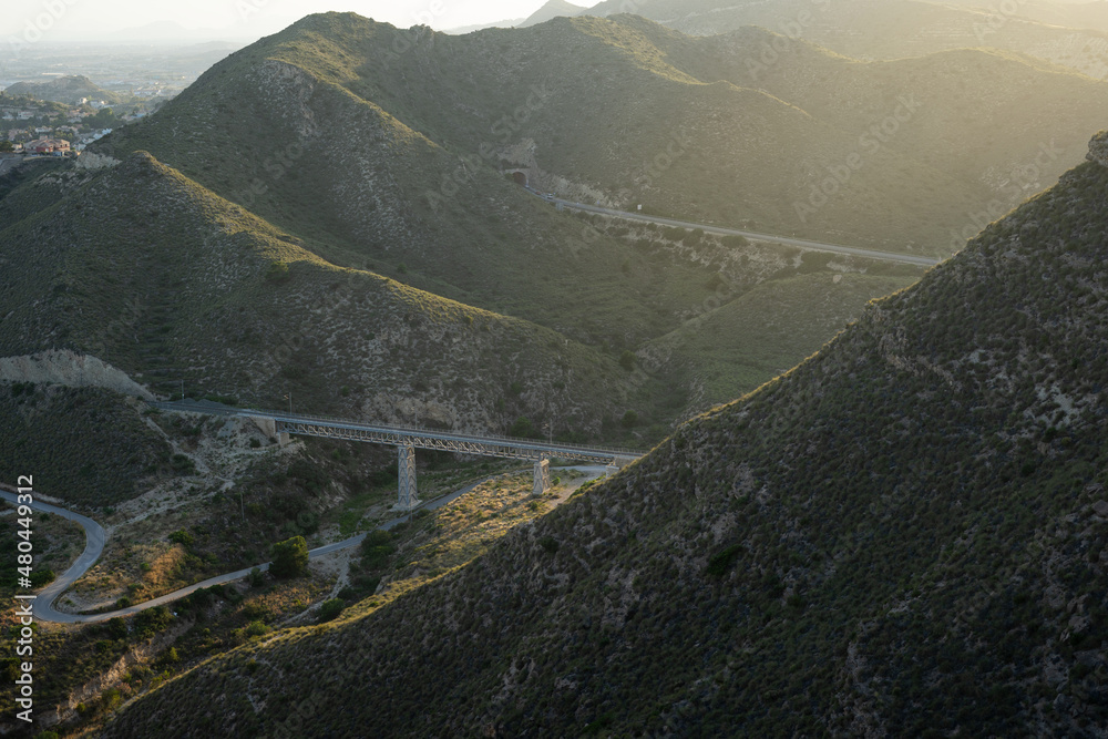 
A large mountain range full of trees in which a bridge can be seen at the bottom during sunset next to the Mediterranean Sea on the Spanish coast.
