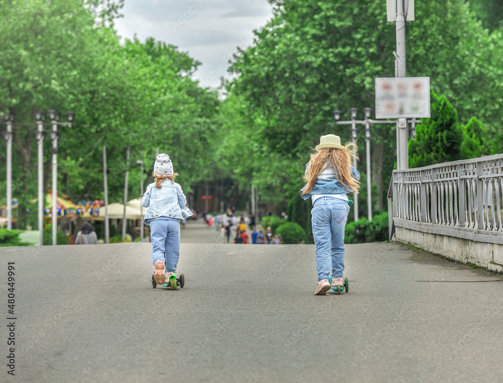 Two sisters on scooters take a walk in a public park, they are fun and active on the high road. Two sisters on scooters take a walk in a public park, they are fun and active on the high road.
