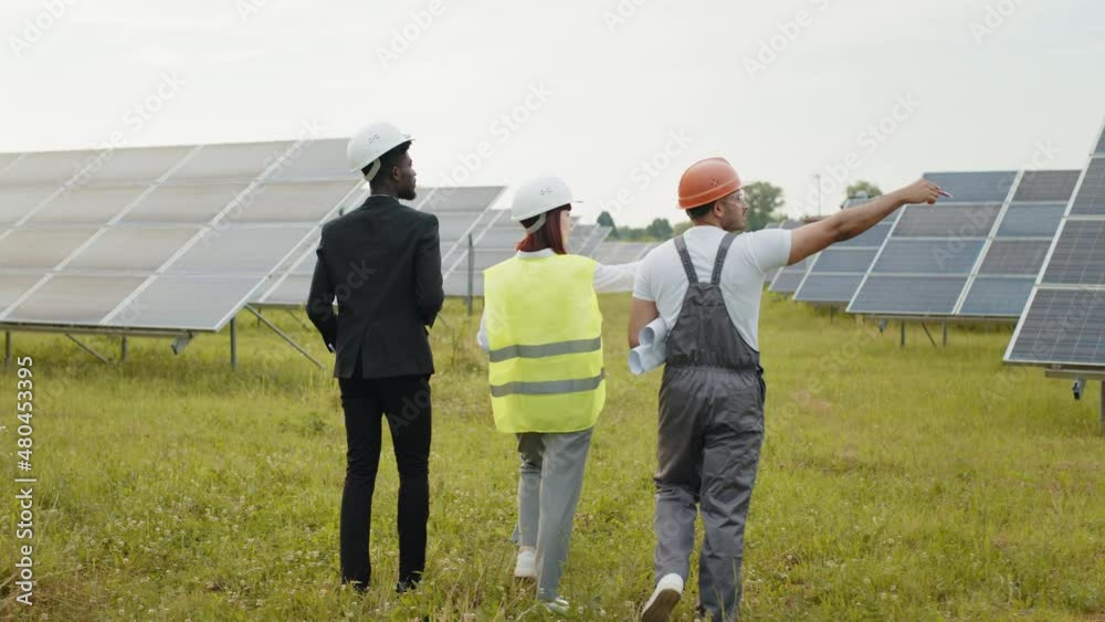 Back view of two inspectors in white helmets walking on solar station with technician in uniform. Multiracial engineers examining work of photovoltaic cells outdoors. Workers walking on solar station. Back view of two inspectors in white helmets walking on solar station with technician in uniform. Multiracial engineers examining work of photovoltaic cells outdoors. Workers walking on solar station.