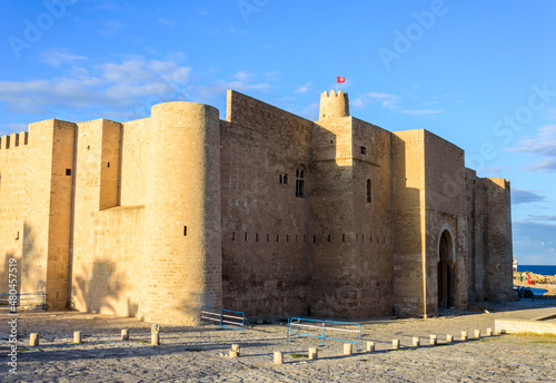 Ancient arabic ribat fortress walls, windows and tower in Monastir, Tunisia, Africa. blue sky, yellow stones, sunlight on the walls, red tunisian in the sky above the tower