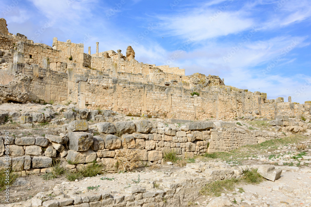 Ruins of the antique Roman city of Thugga in Tunisia, Africa.  Blue sky with clouds, old yellow, grey and brown stone walls and columns, dry yellow grass
