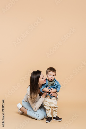 happy mother hugging surprised toddler son on beige.