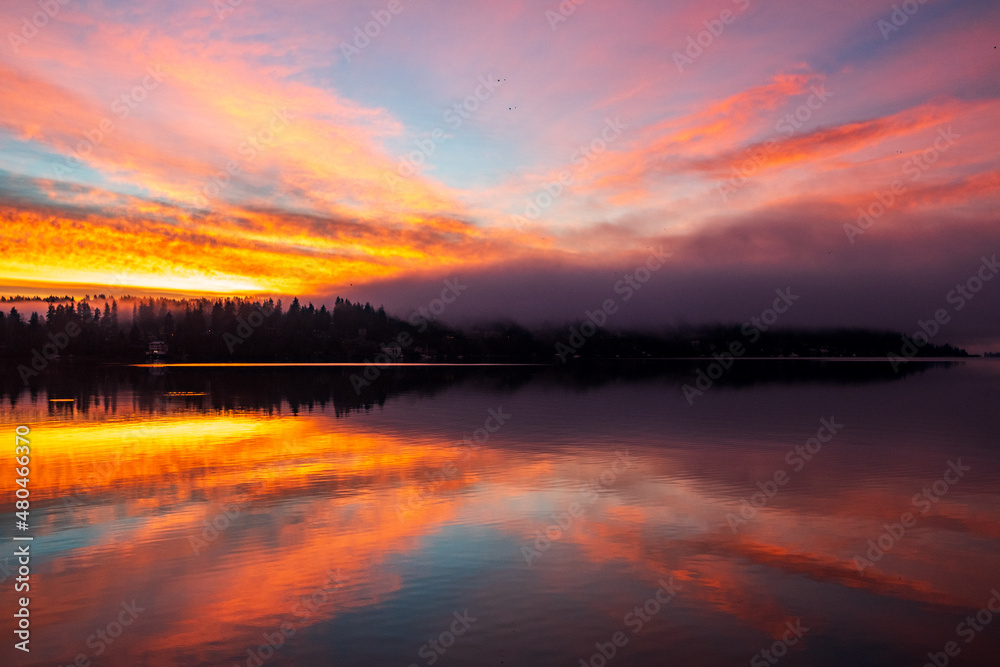 Sunrise over Lake Washington results in a colorful sky and reflection on the water, with foggy forest in the foreground