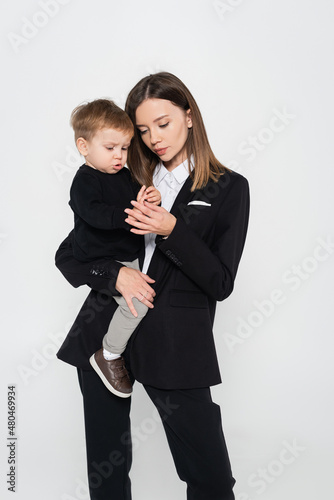 stylish mother in suit holding in arms toddler son isolated on grey.