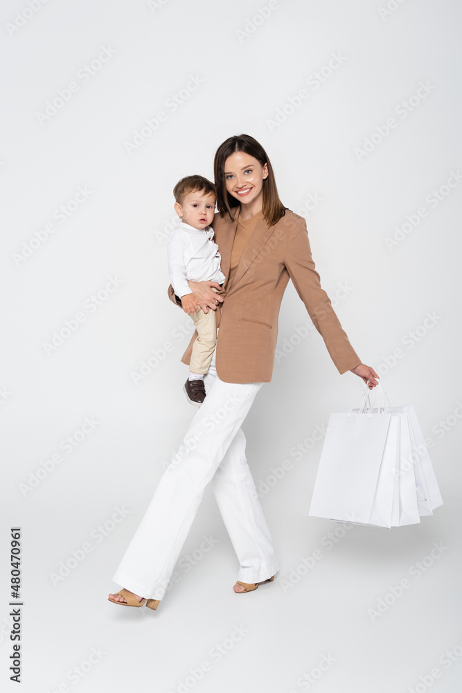 joyful mother holding in arms toddler son and shopping bags while walking on grey. joyful mother holding in arms toddler son and shopping bags while walking on grey.
