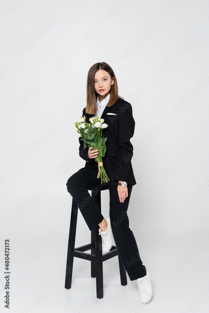 full length of stylish woman in suit holding bouquet of flowers and sitting on chair on grey. full length of stylish woman in suit holding bouquet of flowers and sitting on chair on grey.