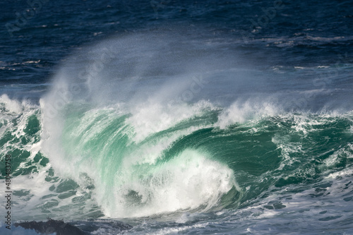 An angry turquoise green color massive rip curl of a wave as it rolls along a beach. The white mist and froth from the wave are foamy and fluffy. The Atlantic Ocean in the background is deep blue. 