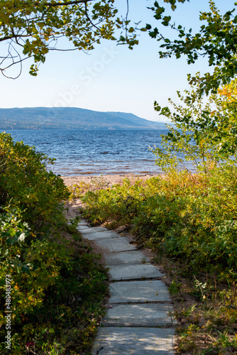 Fototapeta Naklejka Na Ścianę i Meble -  A curved footpath or trail of concrete blocks leading to a sandy beach. The entrance has green and yellow shrubs and trees. There's a mountain in the distance along the riverbank on a sunny day.