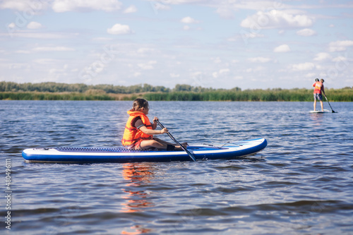 Little Caucasian girl sitting on paddle board alone holding oar in hands and looking at other sup boarders in orange life jacket. Active holidays. Inculcation of love for sports from childhood.