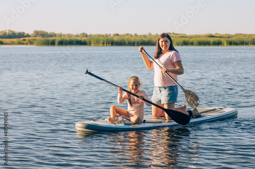 Happy Caucasian mother and little daughter sup boarding with oars in hands looking at camera and smiling on crystal blue lake with green reeds in background. Active lifestyle.