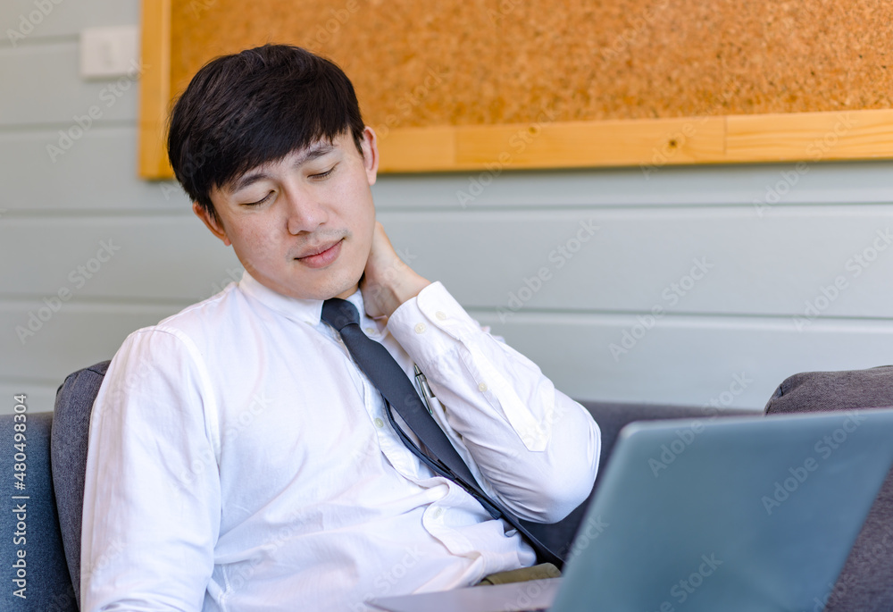 Asian tired overwork male businessman employee in business outfit sitting on sofa working with laptop computer holding hand rubbing massage on stiff injury neck and shoulder from office syndrome Asian tired overwork male businessman employee in business outfit sitting on sofa working with laptop computer holding hand rubbing massage on stiff injury neck and shoulder from office syndrome