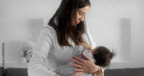 Mother is breastfeeding her newborn baby in the nursery. Mom in a white coat holds and cradles a infant in her arms. Smart watches on the hands of a modern mom. Concept of childhood, parenthood.