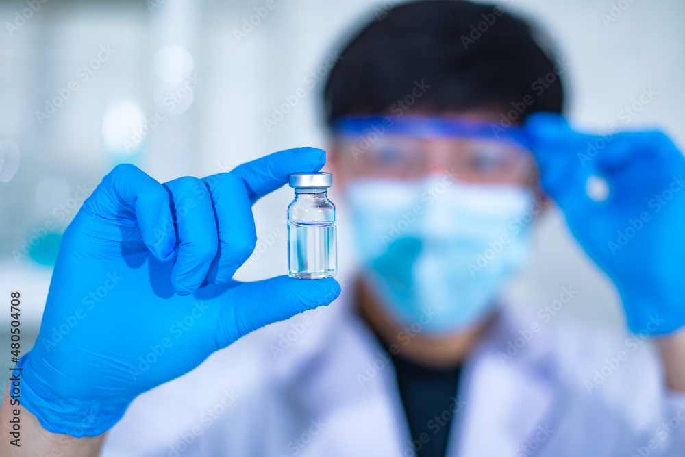 Portrait closeup studio shot of Asian young professional experienced male scientist in white lab coat wearing face mask and safety goggles looking at holding vaccine glass vial in blurred foreground Portrait closeup studio shot of Asian young professional experienced male scientist in white lab coat wearing face mask and safety goggles looking at holding vaccine glass vial in blurred foreground