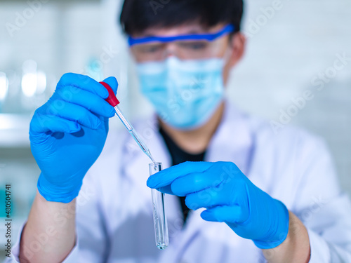 Closeup facial shot of Asian young professional male scientist in white lab coat wearing safety protection goggles glasses using microscope lens looking zooming at microbiology sample on glass plate