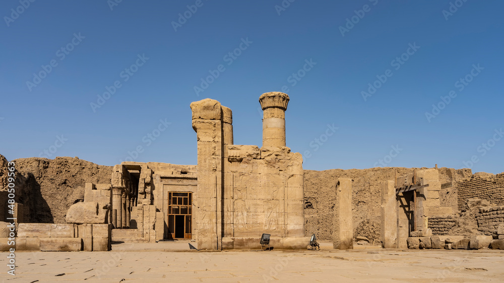 Ruins of the ancient temple of Horus in Edfu. Carvings of gods are visible on the dilapidated walls. Columns with capitals against a clear blue sky. Egypt