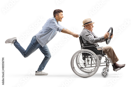 Full length profile shot of a young man running and pushing an elderly man sitting in a wheelchair and holding a steering wheel