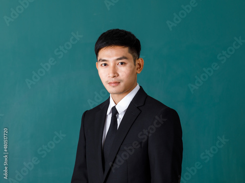 Portrait studio shot of Asian young professional successful male teacher in black formal suit and necktie standing smiling look at camera in front green blackboard background in classroom at school © Bangkok Click Studio