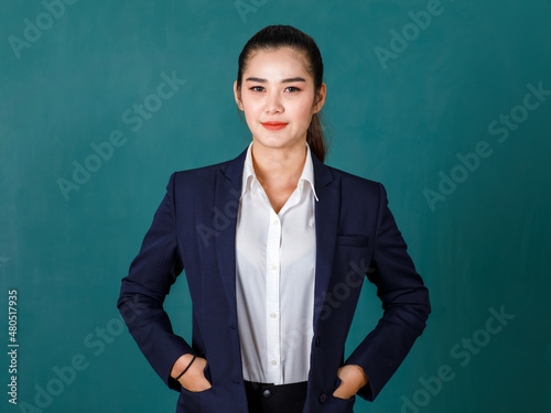 Portrait studio shot of Asian professional ponytail hair female teacher or college student in formal suit standing smiling look at camera hold hands in jacket pockets on green chalkboard background