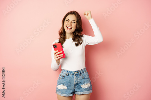 Enthusiastic brunette girl winning on mobile phone, holding smartphone and rejoicing, scream in joy, achieve app goal, pink background