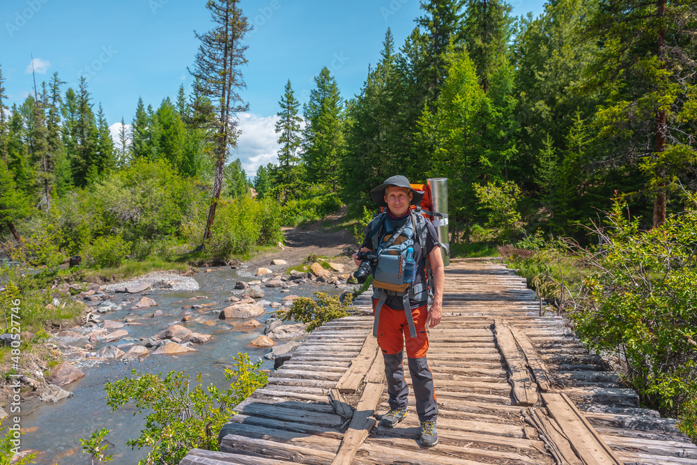 Portrait of traveling man with camera and big backpack in forest on wood bridge through mountain river in bright sun. Vivid landscape with hiker walks in coniferous forest through river in sunny day.