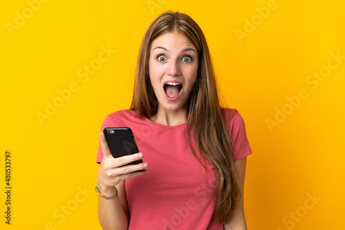 Young woman using mobile phone isolated on yellow background with surprise and shocked facial expression