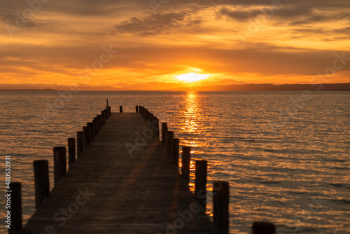 Atmospheric photo of the sunset at Lake Neusiedl with a wooden footbridge running from the picture in the foreground to the left, photographed from an elevated position. The wooden poles cast a shadow